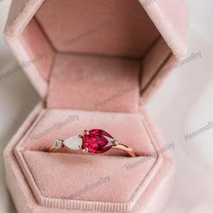 Könnte beinhalten: Ein goldener Ring mit einem birnenförmigen, tiefroten Edelstein und einem kleineren weißen Edelstein. Der Ring befindet sich in einer rosa Samt-Schmuckschatulle. Neben dem weißen Edelstein ist ein kleiner Diamant gesetzt.
