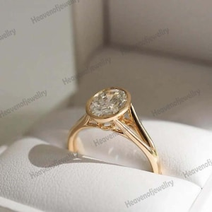 Puede incluir: Un anillo de compromiso de oro con un gran diamante de talla ovalada. El anillo presenta un engaste biselado y un diseño de caña dividida. El anillo se presenta en una caja de joyería blanca, creando contraste.