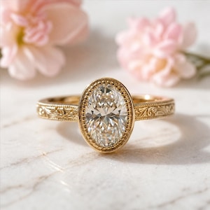 May include: A gold engagement ring with an oval-cut diamond. The ring features a detailed, engraved band and a beaded bezel setting. Pink flowers are softly blurred in the background, adding a touch of color.