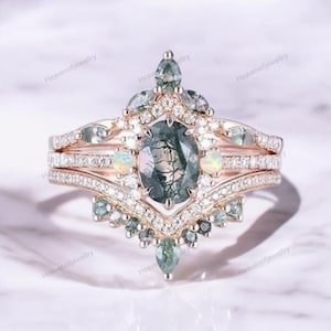 May include: A rose gold ring set with a large oval green moss agate gemstone surrounded by smaller clear gemstones. The ring has a delicate, intricate design with a v-shaped band and a crown-like top.