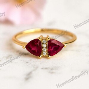 May include: A gold ring featuring two triangular, deep red gemstones flanking a cluster of small, clear stones. The ring's band is slender and polished, with a delicate design. The gemstones are set in a gold setting, creating a striking contrast.