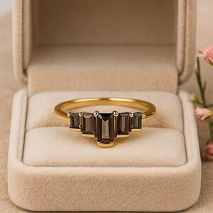 May include: A gold ring with five rectangular, dark brown gemstones set in a row. The ring is displayed in a beige jewelry box, with a soft focus background.