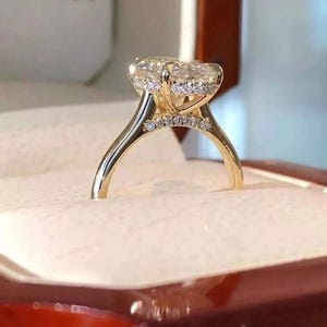 Pode incluir: Um anel de noivado de ouro com um grande diamante central de corte oval. O anel apresenta um halo de diamantes menores ao redor da pedra central e ao longo da banda. O anel é cravejado em uma banda de ouro.