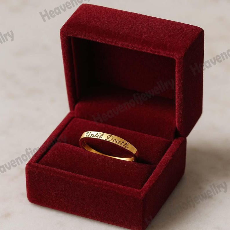 May include: A gold band ring with the inscription "Until Death" inside a red velvet ring box. The ring is centered in the open box, which is a deep red color. The ring's inscription is in a cursive font.