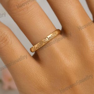May include: A gold-colored ring with the inscription "Until Death" engraved on the band. The ring is shown on a finger, with a soft, neutral background.