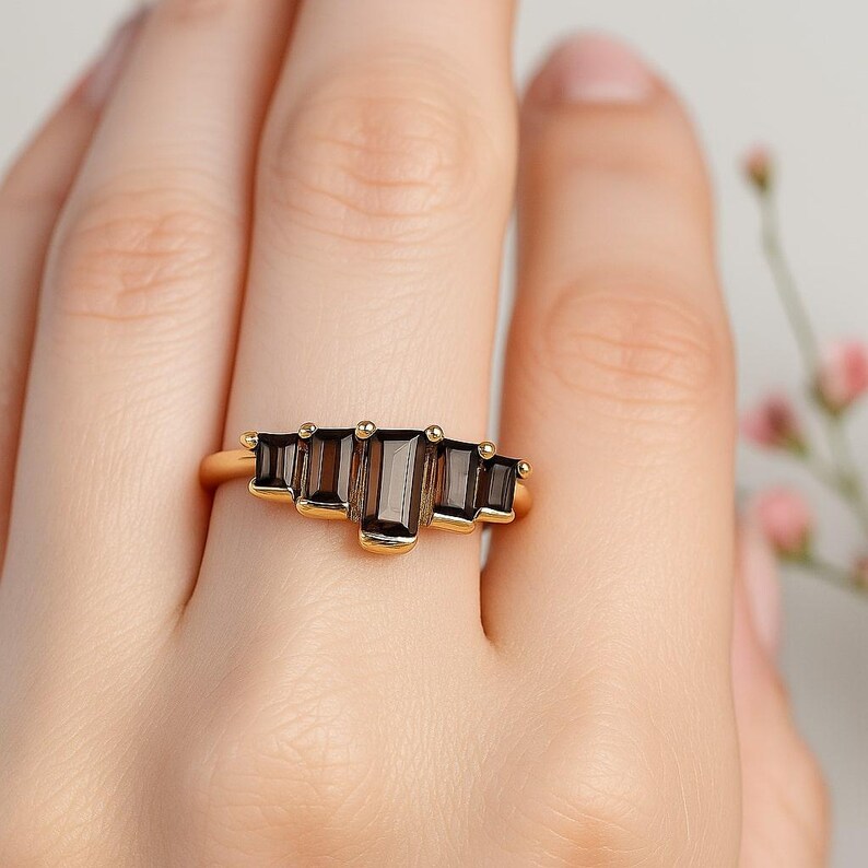 May include: A gold ring featuring five rectangular, dark brown gemstones. The ring's band is a simple, polished gold. The gemstones are arranged in a graduated pattern, with the largest stone in the center.