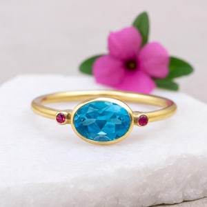 May include: A gold ring featuring a large, oval-cut, bright blue gemstone as the centerpiece, flanked by two smaller, round, red gemstones. The ring is set against a white surface, with a pink flower and green leaves in the background.