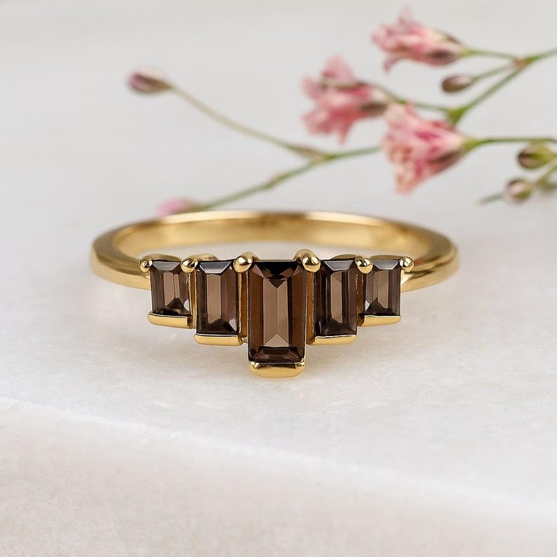 May include: A gold ring featuring five rectangular, smoky brown gemstones. The gemstones are set in a row, with the center stone slightly larger. The ring is set against a white marble surface with pink flowers in the background.