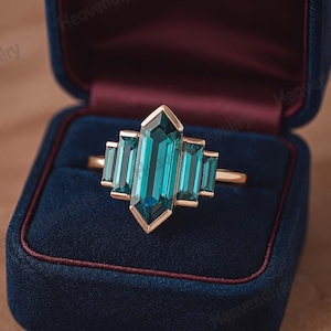 Può includere: Un anello d'oro con una grande pietra preziosa centrale color teal taglio smeraldo, affiancata da quattro pietre rettangolari teal più piccole. L'anello è incastonato in una fascia d'oro e presentato in un cofanetto di velluto blu. Design art déco.
