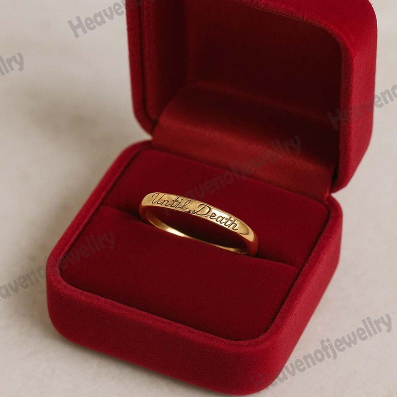 May include: A gold ring with the inscription "Until Death" inside a red velvet ring box. The ring is centered in the open box, showcasing the engraved text. The box is a deep red color.