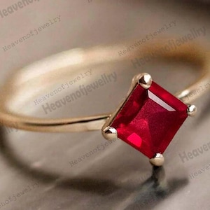 May include: A gold ring with a striking, square-cut, deep red gemstone. The ring has a slender, polished gold band, and the gemstone is held in place by four small prongs. The jewellery is presented against a neutral backdrop, emphasising the design.