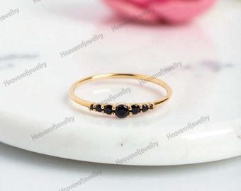 May include: A slender gold ring with five black gemstones set in a row. The ring has a thin, polished band, and the gemstones are closely set, creating a subtle, elegant design. The ring is shown on a white marble surface.