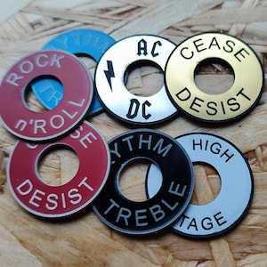 Könnte beinhalten: Eine Sammlung runder Metallunterlegscheiben für Gitarren-Kippschalter in verschiedenen Farben wie Rot, Blau, Silber, Gold, Schwarz und Weiß. Jede Scheibe ist mit Texten wie ROCK n'ROLL, CEASE DESIST, AC DC, RHYTHM TREBLE und HIGH TAGE graviert.