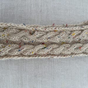 Könnte beinhalten: Ein cremefarbenes Stirnband mit Zopfmuster. Das Stirnband ist mit kleinen roten, gelben und blauen Flecken versehen. Das Accessoire ist aus dickem Garn gefertigt und für kaltes Wetter geeignet.