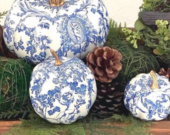 May include: Three white pumpkins decorated with a blue toile pattern featuring cherubs and floral motifs. Each pumpkin has a metallic gold stem. The pumpkins are arranged among green moss, brown pinecones, and small sprigs of greenery.