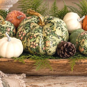 May include: A wooden dough bowl filled with a collection of decorative pumpkins, including solid white and orange gourds alongside several patterned pumpkins featuring green, pink, and gold floral motifs, accented by pinecones and green fern sprigs.
