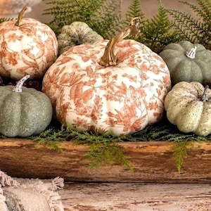 May include: A collection of decorative pumpkins in a wooden dough bowl. The pumpkins feature a mix of solid muted green, speckled light green, and white surfaces with a copper-colored floral pattern. Brown pinecones and green fern sprigs are included.