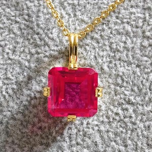 Op de afbeelding: Een goudkleurige ketting met een grote, vierkante, felroze edelsteen hanger. De hanger is gezet in een gouden frame met kleine gouden accenten op elke hoek. De ketting is ook goudkleurig.