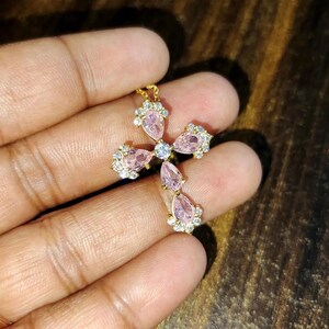 May include: A gold cross pendant necklace with pink pear-shaped crystals. The cross is adorned with smaller clear crystals.