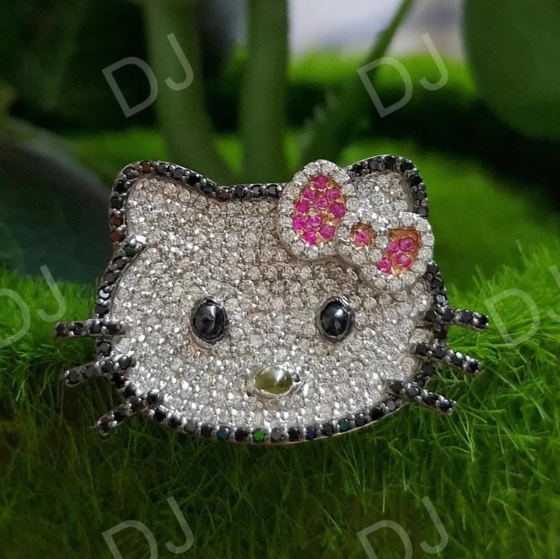 May include: A sparkling, silver-colored brooch in the shape of a cartoon cat's head. The brooch features black eyes, a pink and white bow, and black accents around the edges. The cat's face is covered in small, glittering stones.