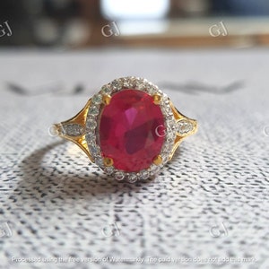 May include: A gold ring featuring a large oval-cut ruby center stone, surrounded by a halo of small, clear gemstones. The ring's band has a split design with additional small gemstones. The ring is set against a neutral background.