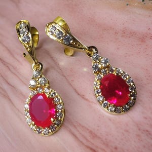 May include: A pair of gold-coloured drop earrings. Each earring has a large, oval-shaped, red gemstone surrounded by a halo of smaller, clear gemstones. The top section of the earrings also has clear gemstones.