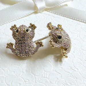 Könnte beinhalten: Ein Paar goldfarbene Frosch-Ohrstecker. Jeder Frosch ist mit kleinen, hellrosa Edelsteinen mit schwarzen Augen besetzt. Die Ohrringe sind vor einem weißen Hintergrund platziert.