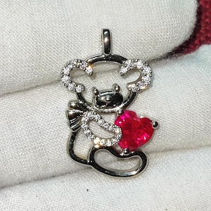 May include: Silver charm pendant featuring a cartoon bear holding a red heart. The bear is outlined with clear sparkling stones.