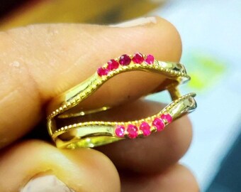 May include: A gold-colored ring with two bands, each lined with small, vibrant red gemstones. The ring is held in a hand, showcasing its intricate design and polished finish. The gemstones are evenly spaced along the bands.