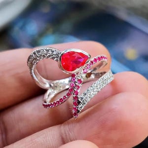 Può includere: Anello d'argento con una gemma rossa brillante a forma di goccia al centro. L'anello presenta un design a doppia fascia, una bordata di piccole pietre rosse e l'altra con pietre chiare e scintillanti. Un gioiello unico.