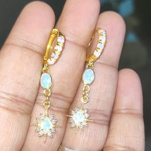 Puede incluir: Un par de pendientes de aro de oro con un colgante de estrella. Los aros están adornados con pequeñas piedras de ópalo blanco. El colgante de estrella tiene un centro de ópalo blanco y está rodeado de piedras transparentes más pequeñas.