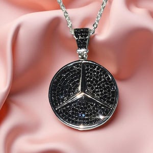May include: A silver pendant necklace with a circular pendant. The pendant features a black, gem-studded Mercedes-Benz logo. The necklace hangs from a delicate chain, set against a soft pink background.