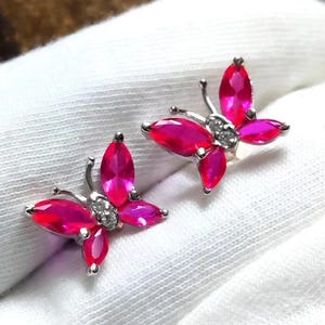 May include: A pair of butterfly-shaped earrings. Each earring features a silver-colored metal frame with marquise-cut, vibrant pink gemstones forming the wings. Small, clear gemstones are set in the center of each butterfly.