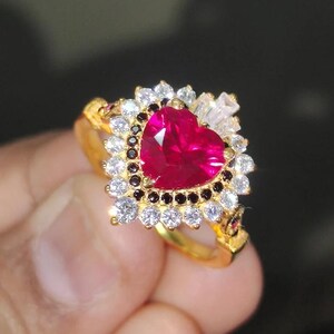 Può includere: Un anello d'oro con un grande rubino a forma di cuore circondato da piccoli diamanti bianchi e neri.