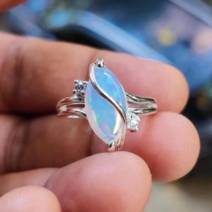 May include: A silver ring featuring a large, oval-shaped opal with a blue and white iridescent sheen. The opal is accented by two smaller, clear gemstones on either side. The ring has a delicate, intertwined band design.