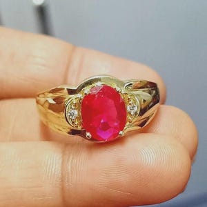 Może przedstawiać: Złoty pierścionek z dużym, owalnym rubinem. Pierścionek ma klasyczny design z dwoma mniejszymi kamieniami akcentującymi po obu stronach rubinu. Pierścionek jest trzymany w dłoni.