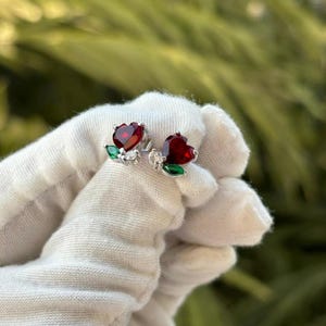 Puede incluir: Un par de pendientes de plata con piedras rojas en forma de corazón y detalles verdes. Los pendientes están diseñados para parecer flores.
