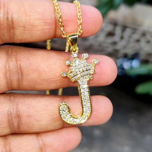 May include: Gold-toned necklace with a pendant shaped like the letter "J" topped with a crown. The pendant and crown are encrusted with small, sparkling crystals. The chain is a delicate link style.