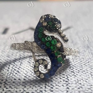 Puede incluir: Un anillo de plata con un diseño de caballito de mar con piedras preciosas azules, verdes y negras. El caballito de mar está rodeado por una banda de piedras blancas más pequeñas.