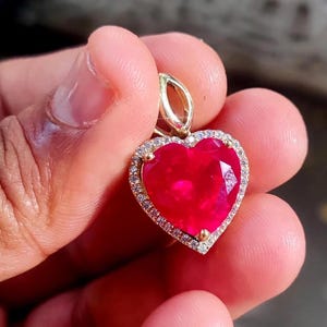 Puede incluir: Un colgante en forma de corazón con una vibrante gema roja en el centro, rodeada por un halo de pequeños diamantes brillantes. El colgante está engastado en un metal dorado. Diseño elegante.