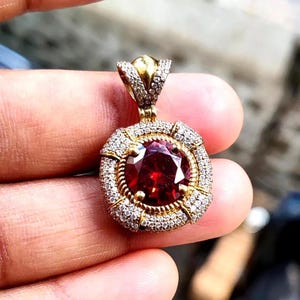 Peut inclure: Un pendentif en or avec une grosse pierre précieuse rouge foncé et ronde au centre. La pierre précieuse est entourée d'un halo de petites pierres précieuses claires. Le pendentif a une bélière dorée décorative.