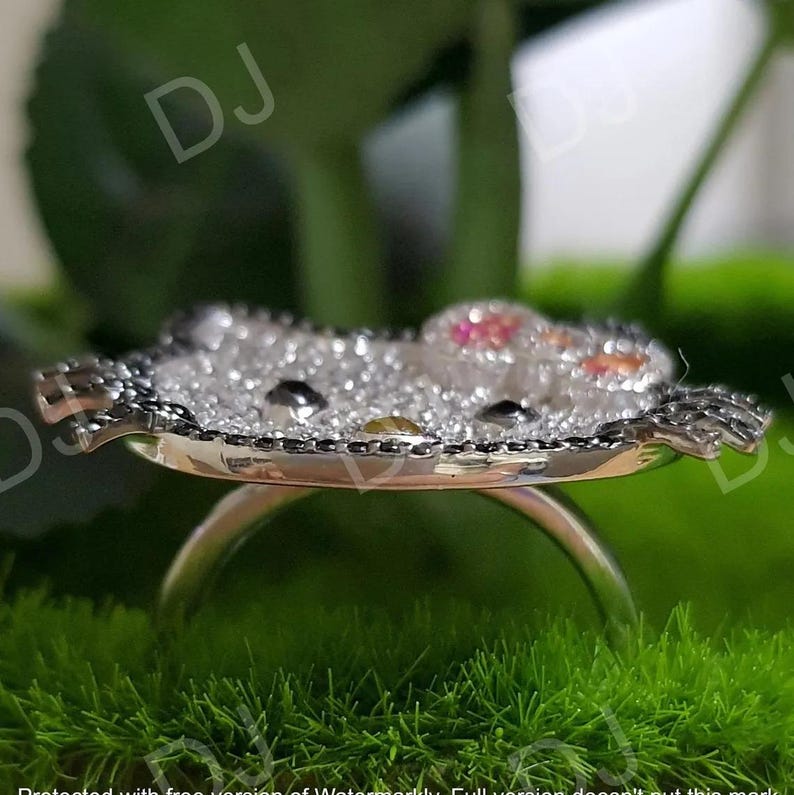 May include: A silver ring featuring a character design with a face made of black and yellow gems. The character's head is encrusted with clear crystals and outlined with black gems. The ring is set against a green background.