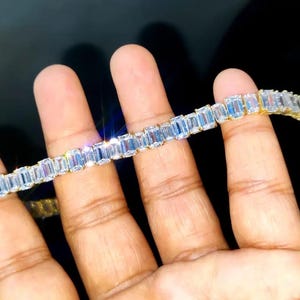 May include: A gold bracelet with a series of rectangular, clear gemstones. The gemstones are set closely together, creating a continuous, sparkling band. The bracelet is held against a dark background, highlighting its brilliance.