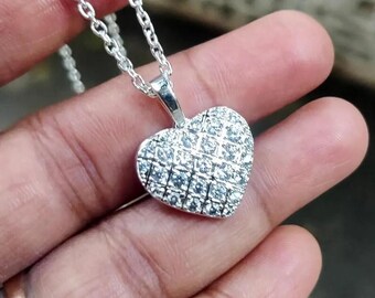 May include: A silver necklace with a heart-shaped pendant. The pendant is covered in small, sparkling stones, and the chain is delicate. The necklace is displayed against a blurred background.