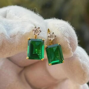May include: A pair of gold-toned earrings featuring large, emerald-green rectangular stones. Each earring has a small, round, clear stone at the top. The earrings are held against a white background.