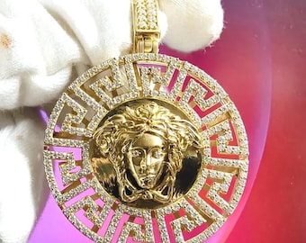 May include: A gold pendant with a Medusa head design surrounded by a circular Greek key pattern, set with clear cubic zirconia stones.