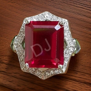 Può includere: Un anello d'argento con una grande pietra preziosa rossa a taglio smeraldo circondata da pietre preziose più piccole e chiare. L'anello ha una montatura decorativa e ornata.