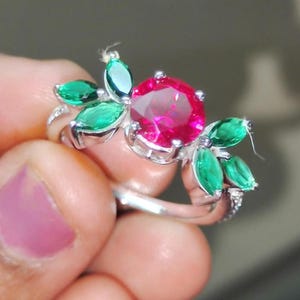 May include: A silver ring featuring a large, round, ruby-red gemstone at the center, surrounded by green marquise-cut stones. The ring's design resembles a flower with a cluster of green leaves.