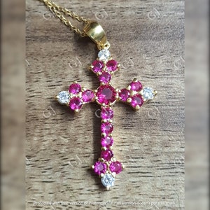 Może przedstawiać: Złoty naszyjnik z wisiorkiem w kształcie krzyża. Krzyż jest ozdobiony licznymi różowymi kamieniami szlachetnymi i kilkoma białymi kamieniami. Naszyjnik jest prezentowany na drewnianej powierzchni.