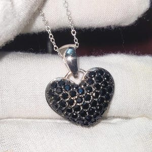 Puede incluir: Un collar de plata con un colgante de corazón negro cubierto de pequeñas piedras negras.